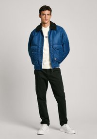 Chaqueta bomber azul con cuello negro, dos bolsillos frontales, usada sobre un sudadera blanca y combinada con pantalones negros y zapatillas blancas.