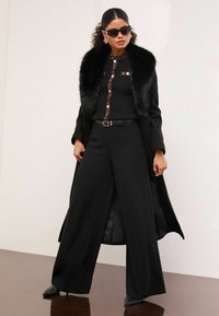 Mustat leveälahkeiset housut, joissa on yhteensopiva pitkä takki, jossa on tekokarvareunus. Yläosassa on ribbikuviointi ja koristeelliset napit.