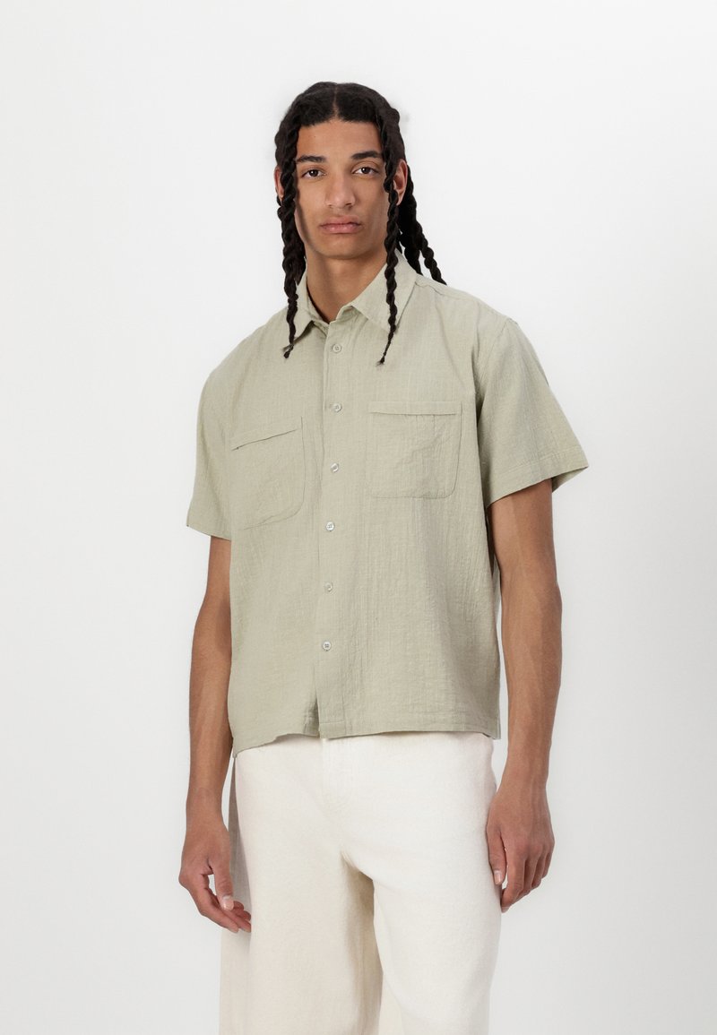 Camicia tessuta a maniche corte, di colore verde chiaro, con due tasche sul petto, chiusura con bottoni e finitura testurizzata. Abbinata a pantaloni color crema.