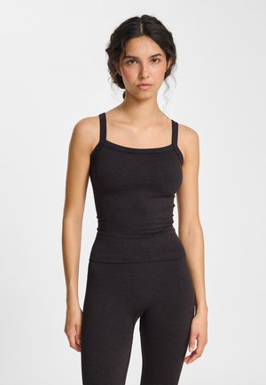 Jonge vrouw met donker krullend haar, gekleed in een nauwsluitend zwart geribd tanktopje en bijpassende legging, staand voor een effen witte achtergrond.