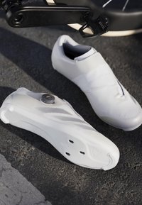 Scarpe da ciclismo bianche con un design elegante, caratterizzate da una parte superiore testurizzata, una rotella di chiusura circolare e una suola in gomma liscia con fori per le tacchette.