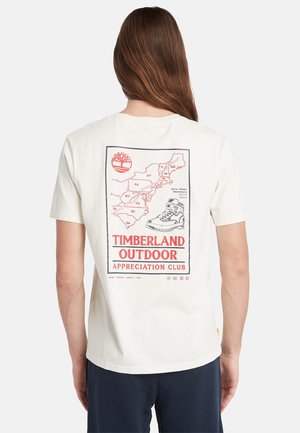 Osoba z długimi brązowymi włosami, nosząca białą koszulkę Timberland, pokazująca na plecach czerwoną i czarną buta do wędrówek oraz mapę wschodnich stanów USA.