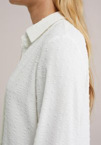 Blusa bianca in tessuto testurizzato con piccoli puntini in rilievo, colletto alla francese e maniche lunghe. Il design è semplice ed elegante.