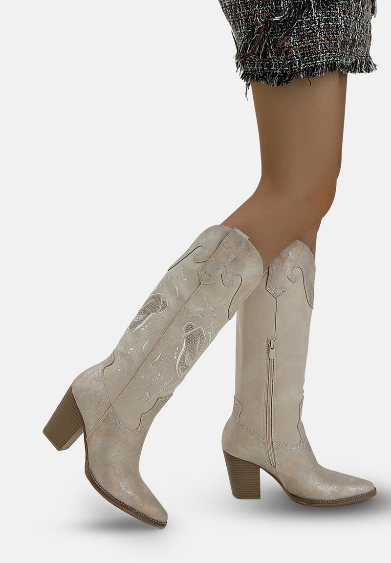 Gambe con stivali al ginocchio in camoscio beige, con tacco a blocco, cerniere laterali e impunture decorative, abbinati a una gonna in tweed.