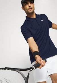 Navyblaues Poloshirt mit Kragen und weißem Nike-Logo, getragen mit weißen Shorts. Die Person hält einen Tennisschläger und trägt ein schwarzes Armband.