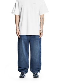 Biała koszulka z krótkim rękawem z subtelnym logo, w połączeniu z luźnymi ciemnoniebieskimi dżinsami i czarnymi butami sportowymi.