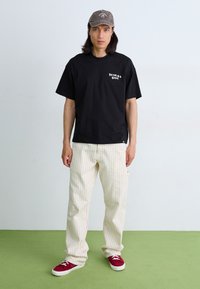 Sort bomuld T-shirt med hvidt logo, parret med cremede stribede bukser. Røde sneakers og en grå kasket fuldender outfittet. Ensfarvet baggrund.