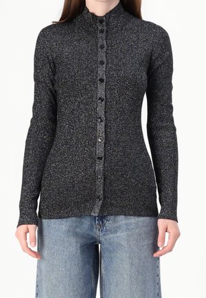 Femme portant un cardigan ajusté noir et argenté scintillant, côtelé, boutonné, à manches longues et un jean en denim bleu clair.