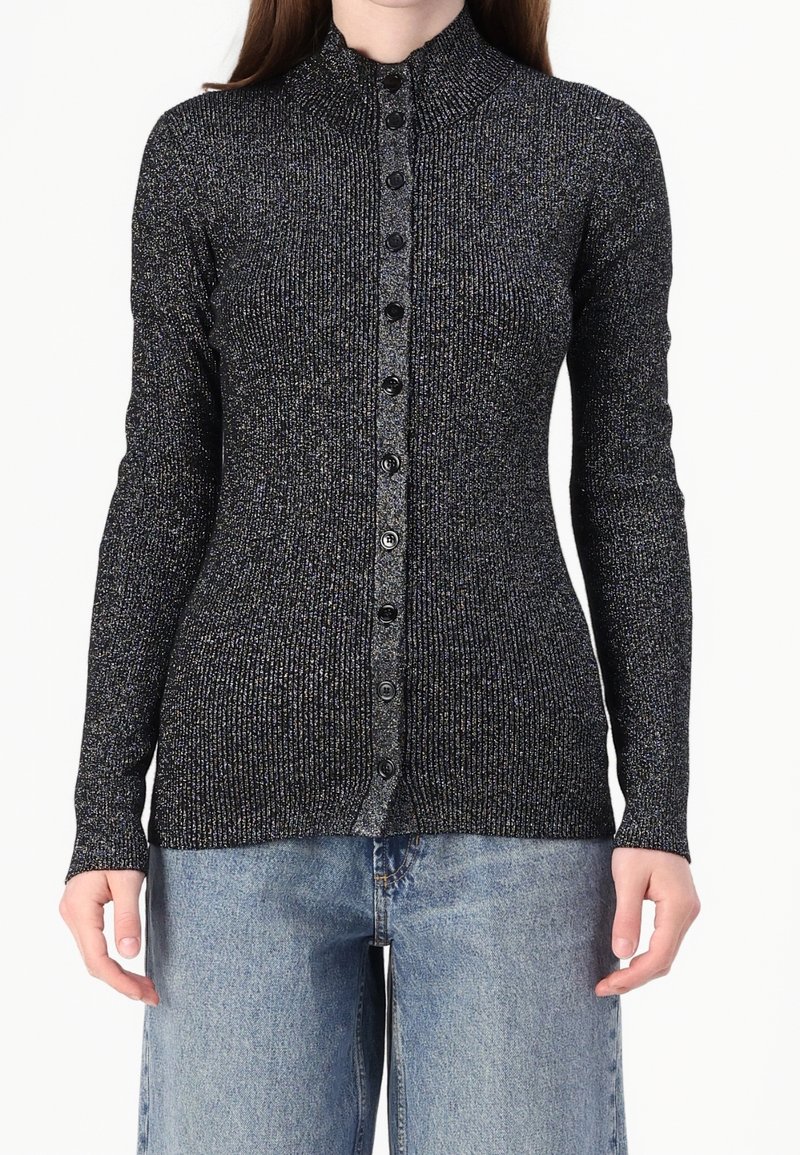 Femme portant un cardigan ajusté noir et argenté scintillant, côtelé, boutonné, à manches longues et un jean en denim bleu clair.