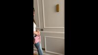 Mujer llevando un bolso rosa, vistiendo una blusa blanca y vaqueros azules, de pie contra una pared beige con paneles decorativos.