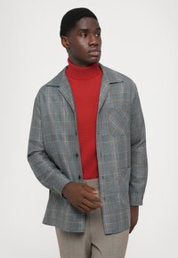 Gray plaid jacket with a notch collar, buttoned front, and one chest pocket, layered over a fitted red turtleneck. Smooth texture.