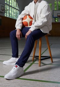 Vit varsityjacka, marinblå byxor och vita sneakers, håller en orange basketboll, sitter på en träpall, i en gymmiljö.