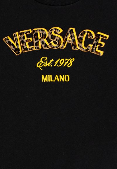 Tissu noir comportant le logo brodé "VERSACE" en jaune avec un motif léopard, "Est. 1978" en jaune, et "MILANO" en jaune vif en dessous.