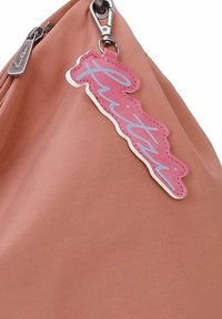 Sac à main doux de couleur terracotta, fabriqué en matériau lisse avec des accessoires en argent. Il est orné d'une étiquette en cuir rose portant le mot "Flutti" en bleu.
