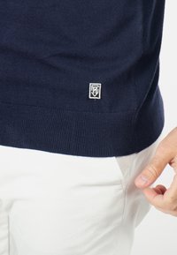 Pull marin avec ourlets et poignets côtelés. Présente une étiquette logo blanche près de l'ourlet. Porté avec un pantalon blanc. Texture de tissu douce.