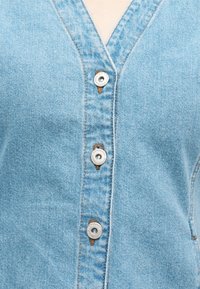 Ανοιχτό μπλε πουκάμισο από denim με τρία ορατά μεταλλικά κουμπιά που είναι κουμπωμένα κατά μήκος μιας κάθετης ραφής στο μπροστινό μέρος.