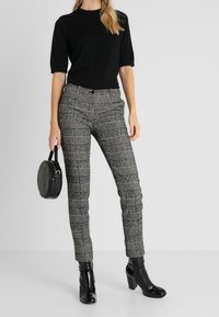 Una blusa negra de manga corta, pantalones de cuadros grises, botas de tacón negras brillantes y un bolso redondo negro acentúan el conjunto.