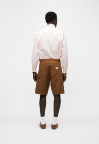 Άνδρας με ανοιχτό ροζ μακρυμάνικο πουκάμισο, καφέ cargo σορτς με πίσω τσέπες, λευκές κάλτσες και καφέ μοκασίνια, στέκεται και κοιτάζει προς τα πίσω.