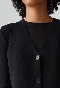 Černá pletená vesta s výstřihem do V, se třemi knoflíkovými zapínáními a měkkou texturou. Nositá přes černý dlouhý rukáv.