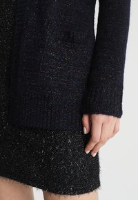 Tmavě námořnicky modrý pletený svetr s třpytivými flíčky, žebrovanými manžetami a boční kapsou, kombinovaný s texturovanou černou minisukní.