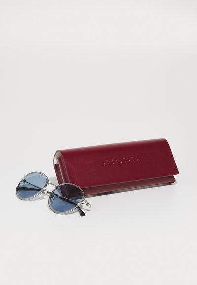 Ochelari de soare Gucci cu lentile rotunde albastre, rame din metal argintiu și o carcasă din piele vișinie embosată cu "Gucci."