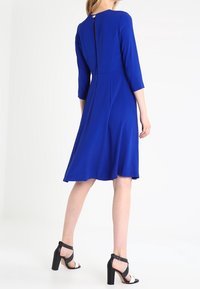 Królewsko niebieska sukienka do kolan z rękawami 3/4, okrągłym dekoltem i ukrytym zamkiem z tyłu. Połączona z czarnymi szpilkami.