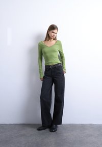 Νεαρή γυναίκα με πράσινο μακρυμάνικο μπλουζάκι, φαρδιά μαύρα τζιν με πλεκτή ζώνη, μαύρα παπούτσια, στέκεται μπροστά σε λευκό τοίχο σε γκρι δάπεδο.