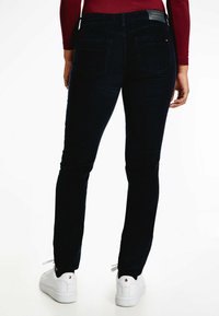 Pantalones de corduroy azul oscuro con un corte ajustado, que cuentan con bolsillos traseros y una etiqueta de marca en la cinturilla, combinados con zapatillas blancas.