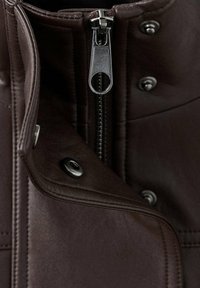 Gros plan d'une veste en cuir marron foncé avec une fermeture éclair argentée et des boutons-pression noirs partiellement ouverts.