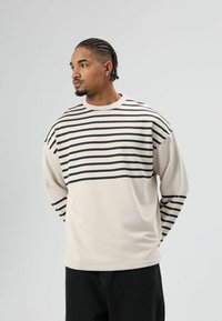 Sweat-shirt beige à manches longues avec des rayures horizontales noires sur le haut de la poitrine et les épaules. Coupe ample, col rond côtelé et manches côtelées.