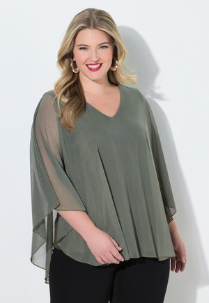 Lächelnde Frau mit langen blonden Haaren, die Creolen trägt und ein lockeres, durchsichtiges olivgrünes Top über schwarzen Hosen trägt, vor weißem Hintergrund stehend.