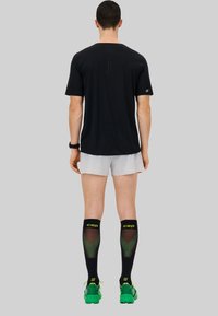 Schwarzes Sport-T-Shirt mit durchbrochenem Design auf dem Rücken, kombiniert mit hellen Shorts und schwarzen Kompressionssocken mit grünen Akzenten.