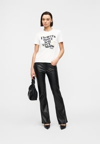 Γυναίκα με λευκό T-shirt με λουλουδάτο κείμενο "Τα λουλούδια δεν μιλούν, δείχνουν", μαύρο παντελόνι από δερματίνη, μαύρες γόβες και κρατώντας μια μαύρη τσάντα.