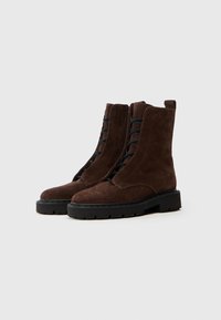 Bottines en suédine marron avec une semelle en caoutchouc noir. Elles disposent d'un laçage à l'avant et d'une fermeture éclair sur le côté pour un enfilage facile. Design plat et épais.