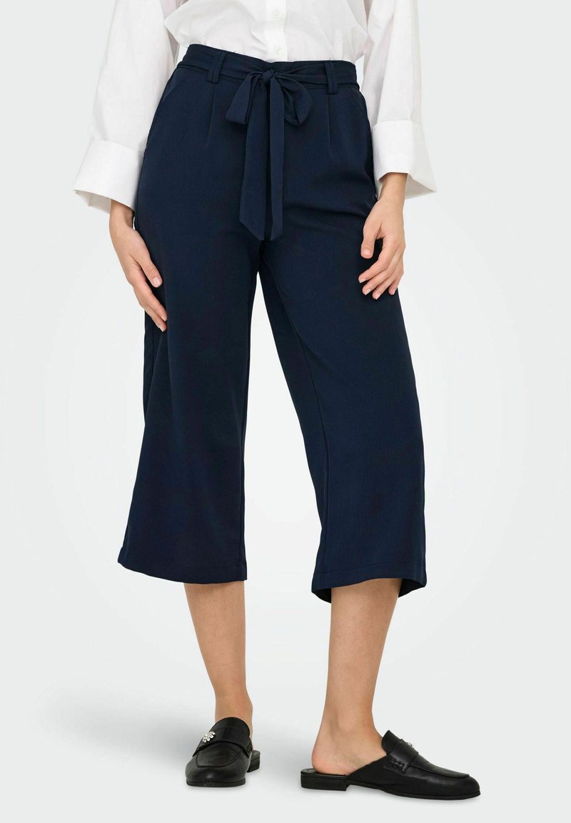 Femme portant un pantalon large cropped bleu marine avec une ceinture nouée à l'avant et des mocassins noirs sans lacets.