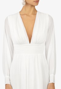Robe blanche à manches longues en tulle, décolleté en V profond et taille froncée. Texture lisse avec une jupe fluide et des accents minimalistes.