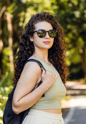 Black sunglasses with round lenses, a light green ribbed crop top, and white loose pants; carrying a black backpack over one shoulder.