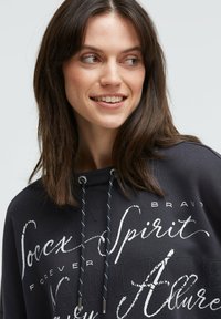 Schwarzer Hoodie mit weißem Text und Designs, ausgestattet mit einem Kapuzenzug. Weiche Textur, lockere Passform und moderner Grafikstil.