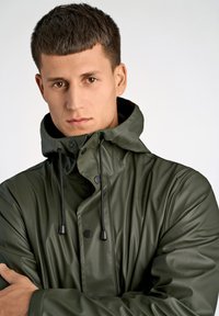 Chaqueta impermeable verde con cuello alto, botones a presión y cordones ajustables. Fabricada en un material brillante y suave con mangas largas.