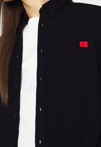 Camisa negra de botones con un parche de logo rojo en el bolsillo del pecho. Hecha de algodón, cuenta con un collar y una camiseta interior blanca en contraste.