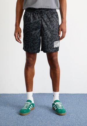 Mężczyzna w czarnych, wzorzystych szortach Adidas z białym logo, białych skarpetach Adidas i zielonych butach sportowych Adidas na niebieskim dywanie.