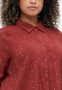 Blouse rouge avec un devant à boutons, un col, deux poches poitrine et des embellissements en pois blancs sur toute sa surface. Tissu doux avec une texture lisse.