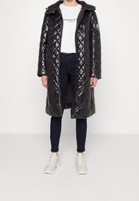 Cappotto lungo nero trapuntato con finitura lucida, dotato di cappuccio e motivo a rombi. Indossato sopra una camicia bianca e pantaloni scuri, con scarpe neutre.