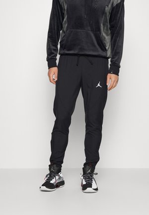 Mand iført sorte Nike Jordan træningsbukser og sort-hvide sneakers står mod en ensfarvet baggrund.