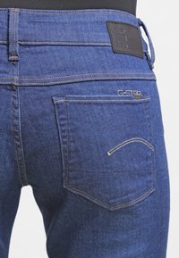 Jeans de mezclilla azul con un primer plano de la vista trasera, que presenta una etiqueta de cuero, bolsillos cosidos y un detalle de costura en contraste.