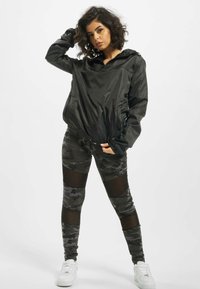 Fekete, könnyű kapucnis dzseki, elülső cipzárral, magasan derekú kamuflázs leggings-szel, amely hálós panelekből áll, és fehér sportcipővel van párosítva.