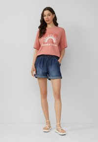 T-shirt oversize rose clair avec un graphique "Golden Hour", assortie à un short en denim foncé avec des ourlets roulés et une ceinture élastique.