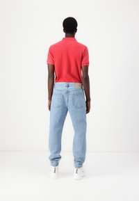 Hombre de pie frente a una pared blanca, vestido con una camiseta polo roja, jeans azul claros y zapatillas blancas con texto negro en el talón.