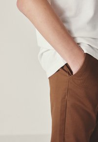 Pantalon en coton marron avec une poche latérale, associé à une chemise blanche. Le tissu est lisse et la coupe est décontractée.