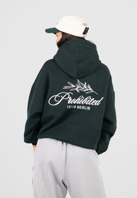 Donna che indossa un felpa verde scuro con la scritta "Prohibited 10119 Berlin" e un grafico di foglia sulla schiena, abbinata a pantaloni grigi chiari e un cappellino bianco.